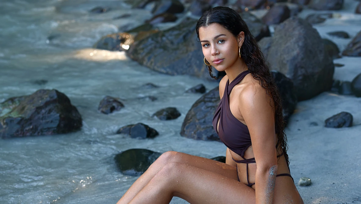 Woman in brown bikini sitting in shallow water surrounded by rocks at dusk.