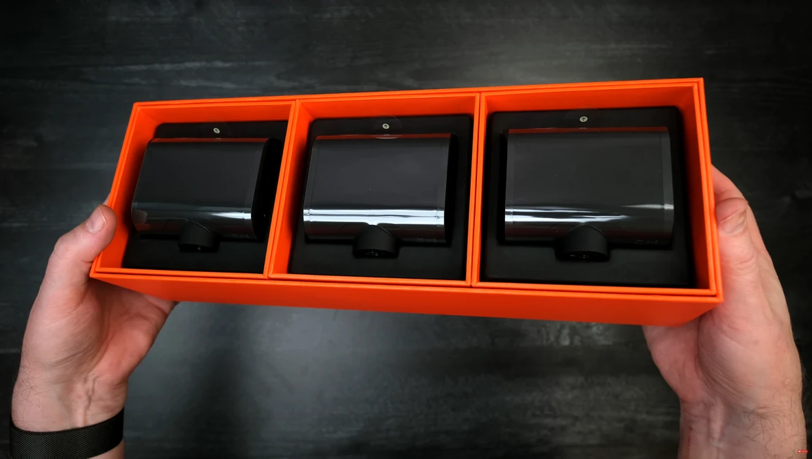 Person holding an orange display case containing three black square lens filters or optical elements.