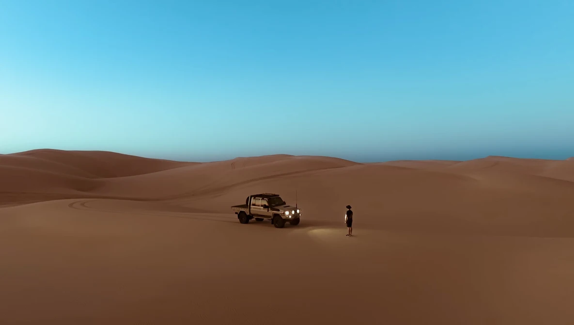 A rugged vehicle and lone figure on vast, undulating sand dunes under clear blue sky.