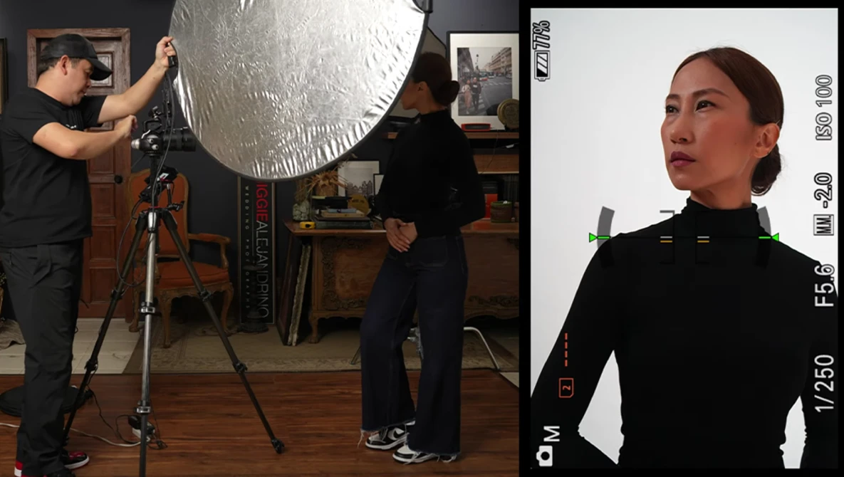Studio portrait setup with large reflector on tripod and subject in black clothing against white background.