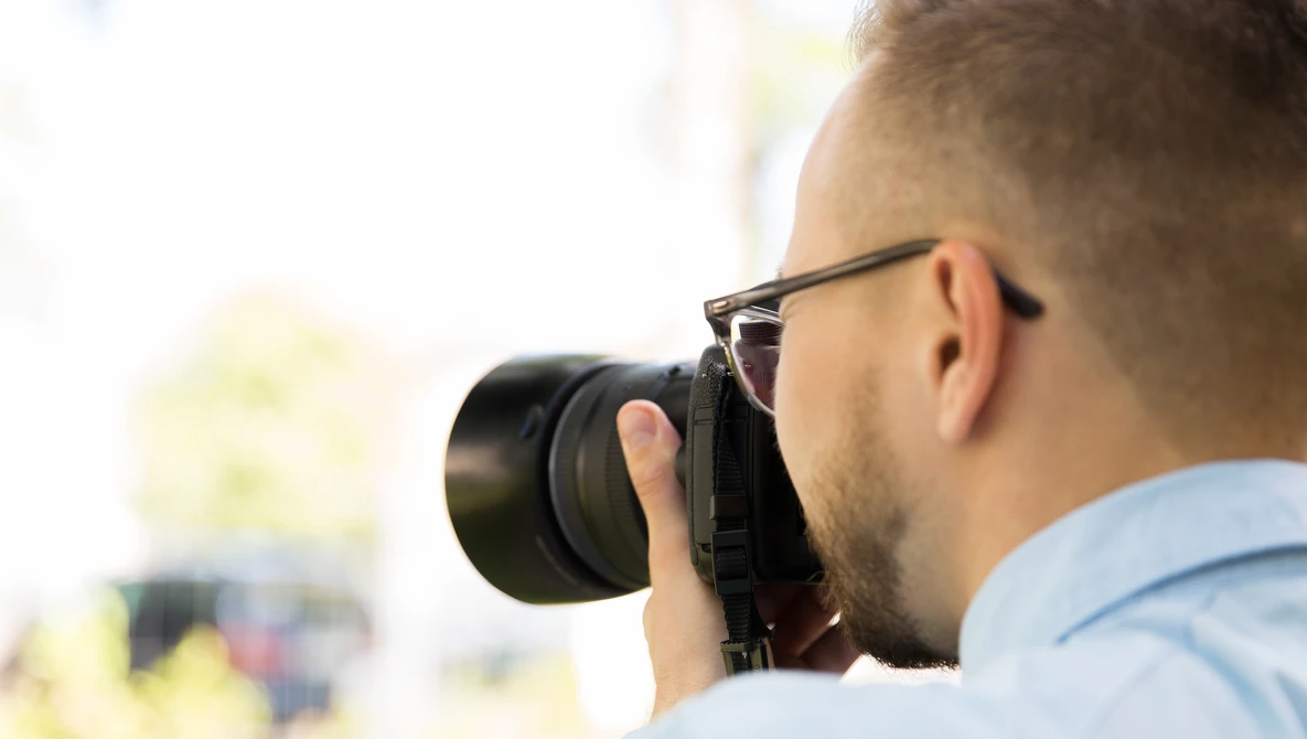 Photographer wearing glasses and light blue shirt holding camera to face while shooting outdoors.