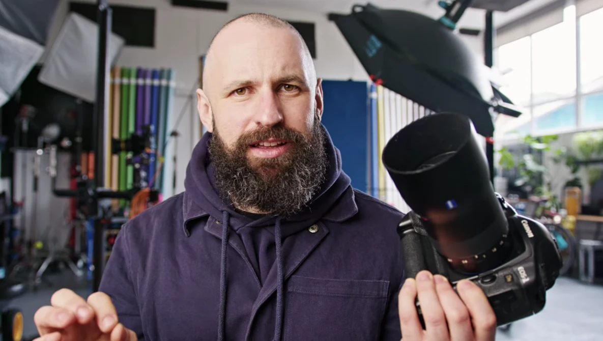 Bearded photographer holding a professional DSLR camera in a studio setting with lighting equipment visible.