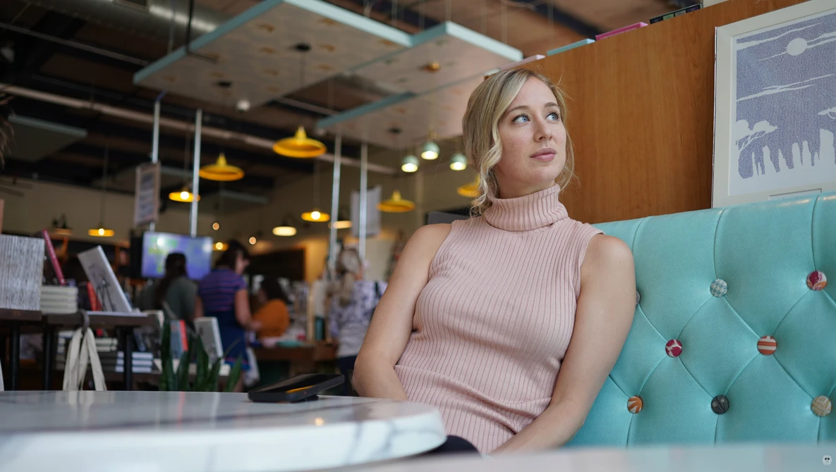 Woman in pink sleeveless turtleneck sitting on turquoise couch in modern office space.