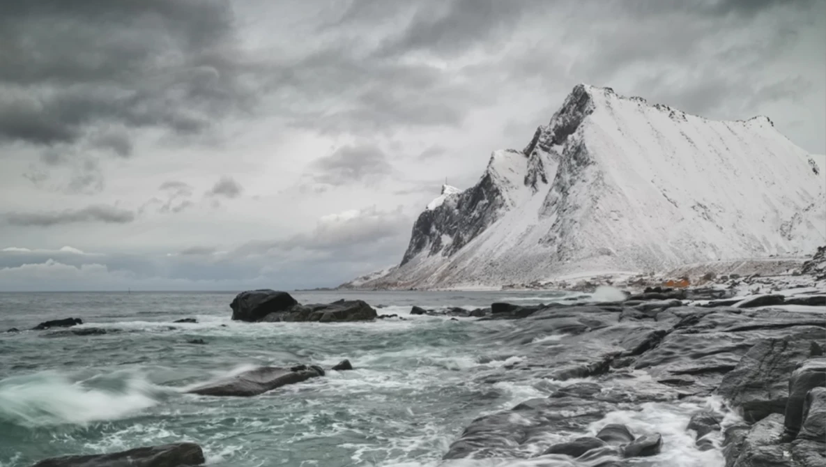 Snow-covered mountain peak rising from rocky shoreline with turbulent sea and overcast sky.