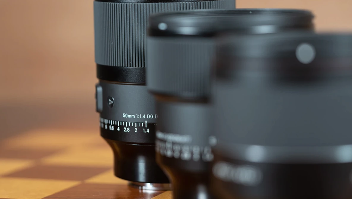 Close-up of a 50mm f/1.4 lens with focus ring and aperture scale visible, with additional lenses blurred in the background.