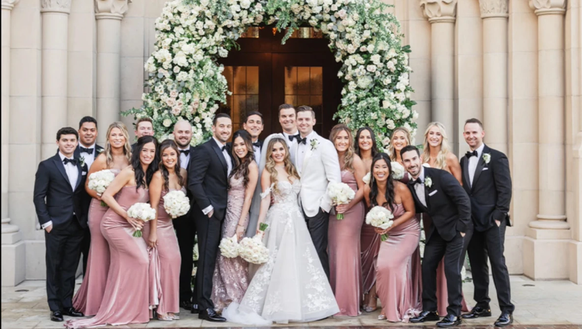 Bride and groom centered with wedding party in formal attire arranged in front of classical columns and floral archway.