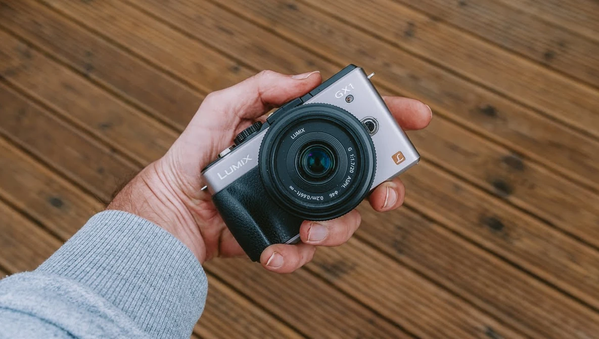 Hand holding a compact mirrorless camera with a fixed lens against a wooden surface.