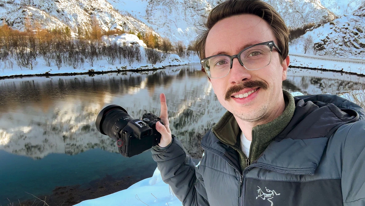 Photographer holding a camera with snow-covered mountains and a frozen river in the background.