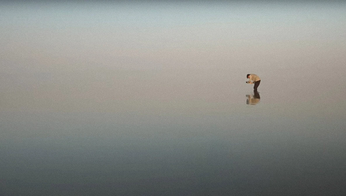 Person standing alone in shallow water with mirror-like reflections under overcast sky.