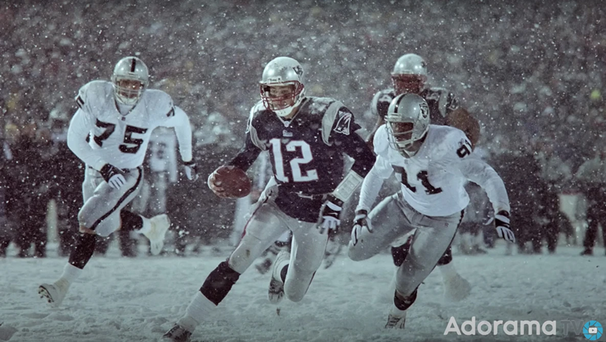 Football players in heavy snow, one in dark uniform carrying the ball while two in white uniforms defend.