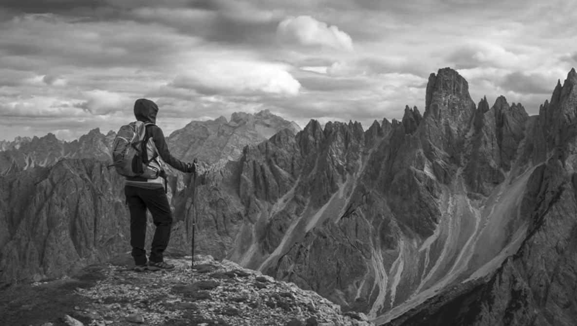 Hiker with backpack and trekking poles standing on a rocky ridge overlooking dramatic jagged peaks.