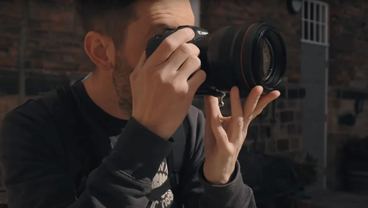Male photographer holding a DSLR camera up to his face while shooting outdoors against a brick building backdrop.