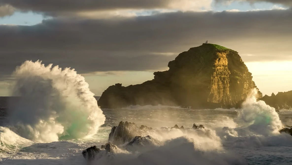 Massive waves crash against a rocky coastal outcrop during golden hour, with dramatic cloudy sky overhead.