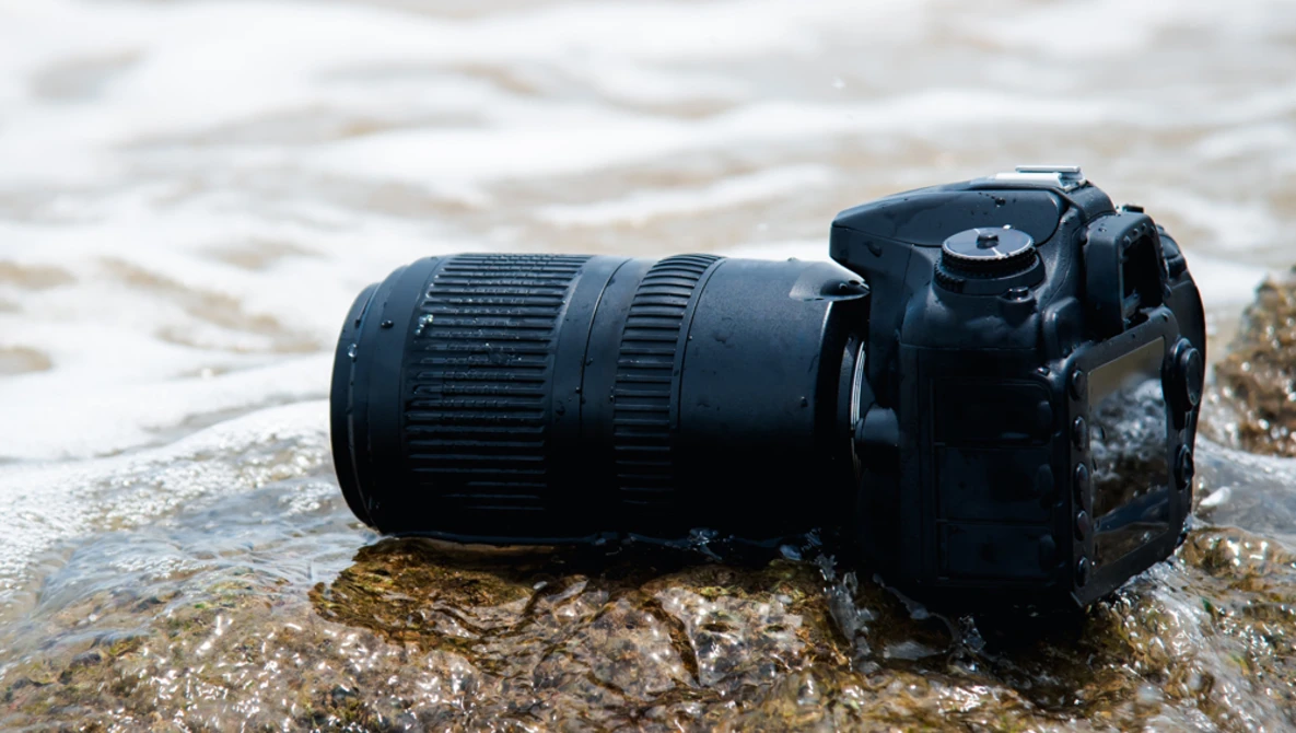 Black DSLR camera with telephoto lens partially submerged in shallow ocean water on a rocky shore.
