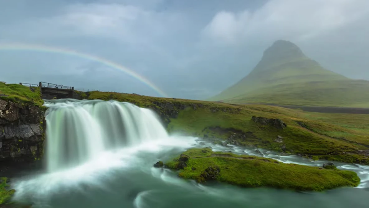 Waterfall cascading into turquoise pool with moss-covered rocks, rainbow visible in mist, mountain peak in background.