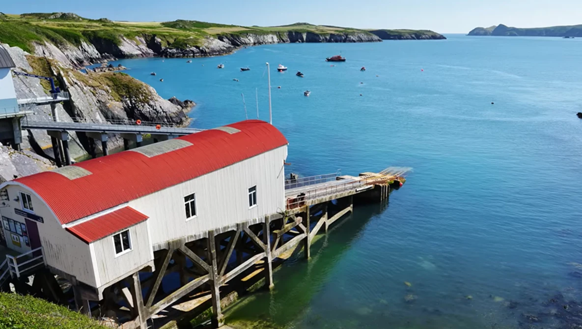 Red-roofed boathouse on wooden stilts overlooking a turquoise cove with white cliffs and moored boats.