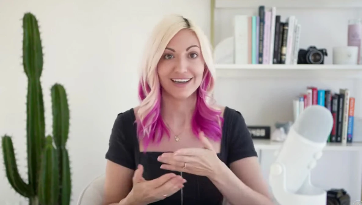 Woman with blonde and pink ombre hair wearing black shirt, gesturing while speaking to camera in bright home office.