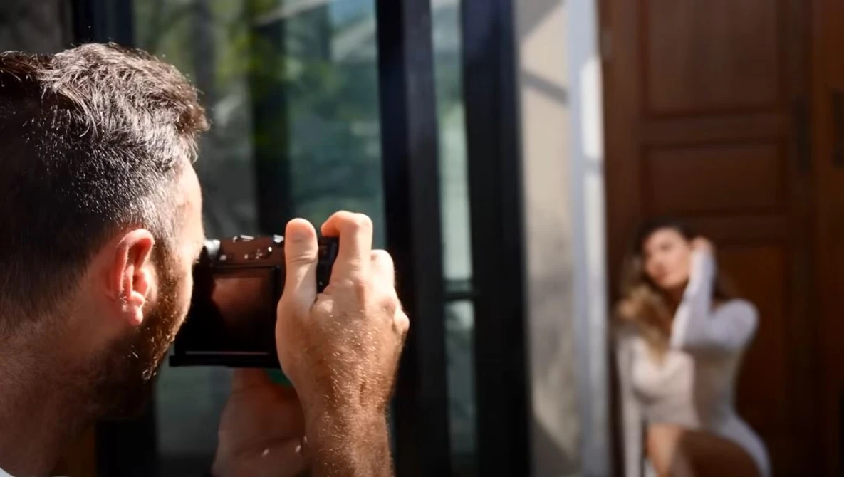 Photographer holding a camera up to his face, capturing an image of a woman visible through a doorway in the background.