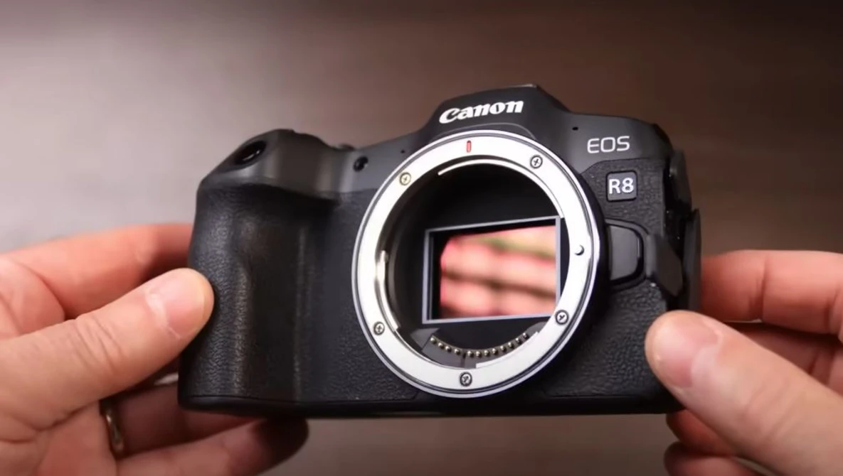 Canon EOS R8 mirrorless camera held in two hands with sensor exposed, shown against a brown gradient background.