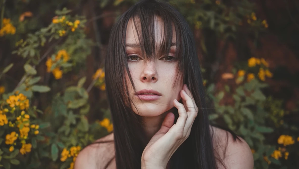 Woman with long dark hair and bangs gazing pensively beside blooming yellow flowers.