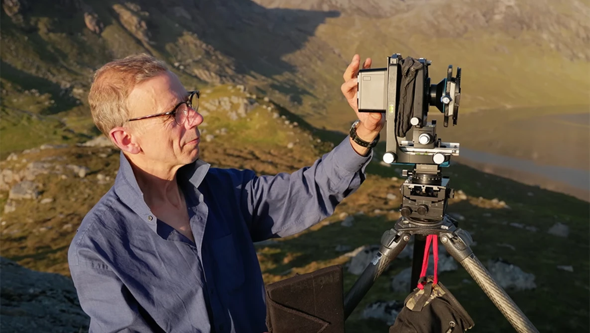 Photographer adjusting a large format camera mounted on a tripod overlooking a mountainous valley landscape.