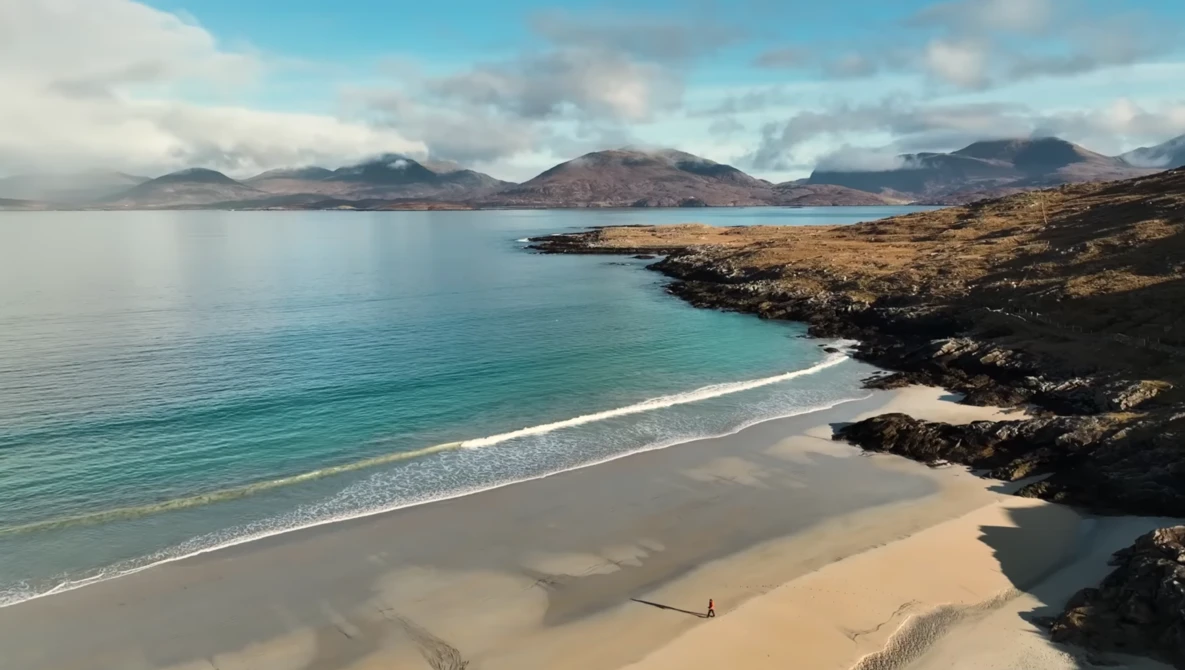 Coastal beach with turquoise water, golden sand, and dark rocky outcrops under blue sky with mountains in distance.