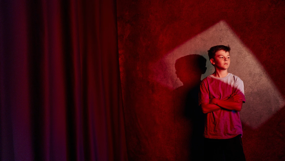Young man in red shirt standing with arms crossed against red wall with dramatic shadow and stage lighting.