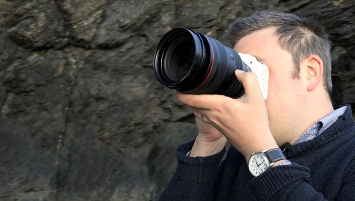 Photographer holding a telephoto lens up to his face against a rough stone wall background.