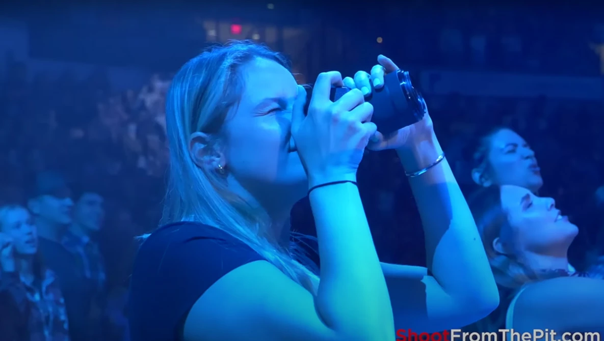 Woman singing into microphone at concert with blue stage lighting and crowd in background.