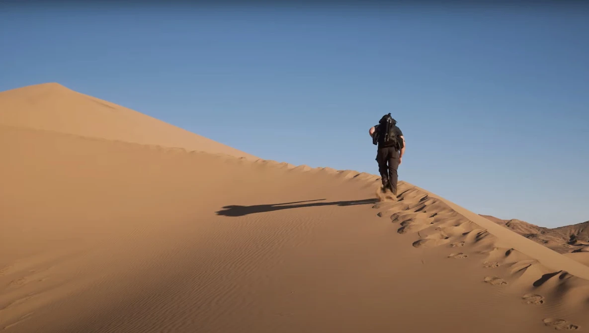 Hiker ascending a steep sand dune under clear blue sky, casting a long shadow on the golden slope.
