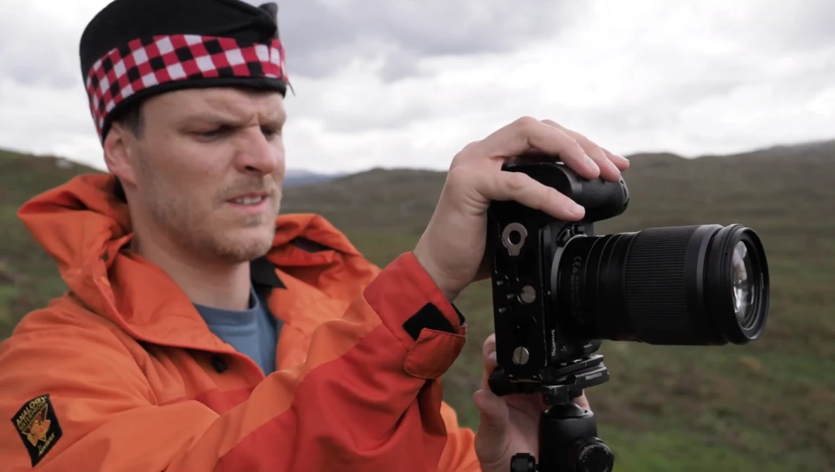 Photographer in orange jacket and red plaid headband holding a DSLR camera with telephoto lens in an open moorland landscape.