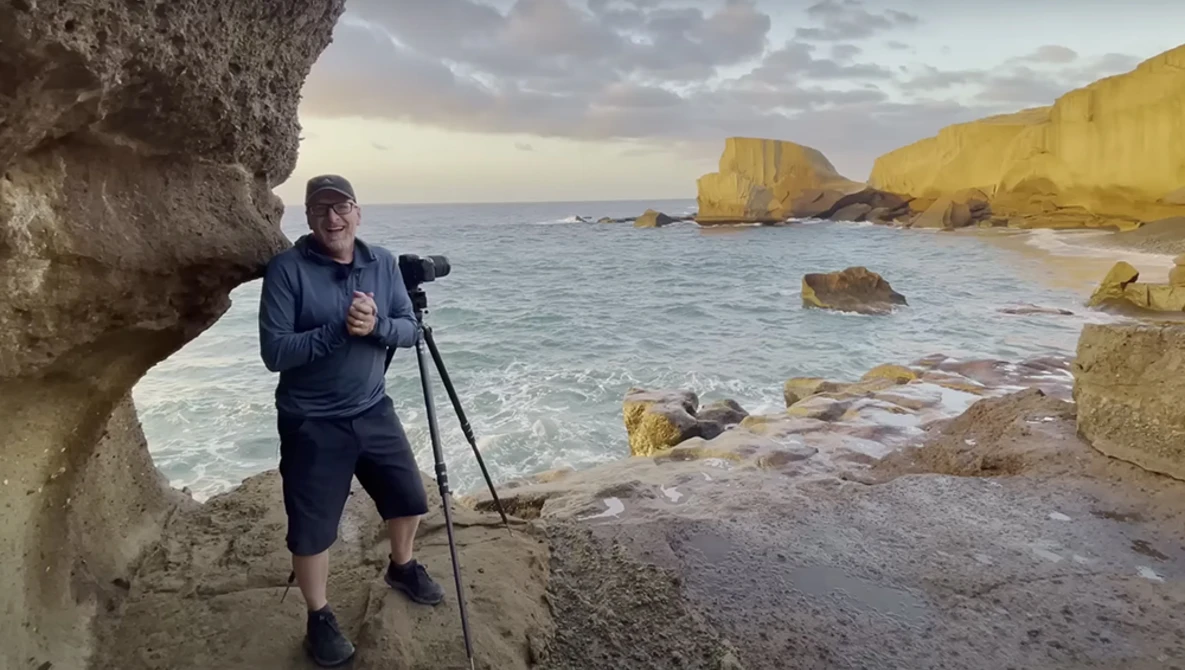 Photographer with tripod-mounted camera shooting coastal rock formations during golden hour.