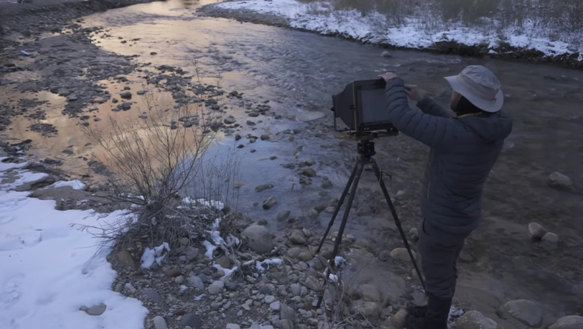 Photographer operating a camera on tripod at the edge of a rocky shoreline with incoming waves.
