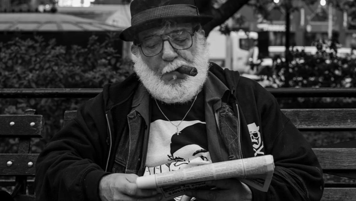Older man with white beard wearing glasses and a hat, smoking a cigar while holding a newspaper on a park bench.