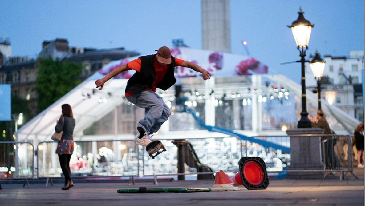 Skateboarder performing a trick mid-air above a rail with an urban fountain and cityscape in the background.