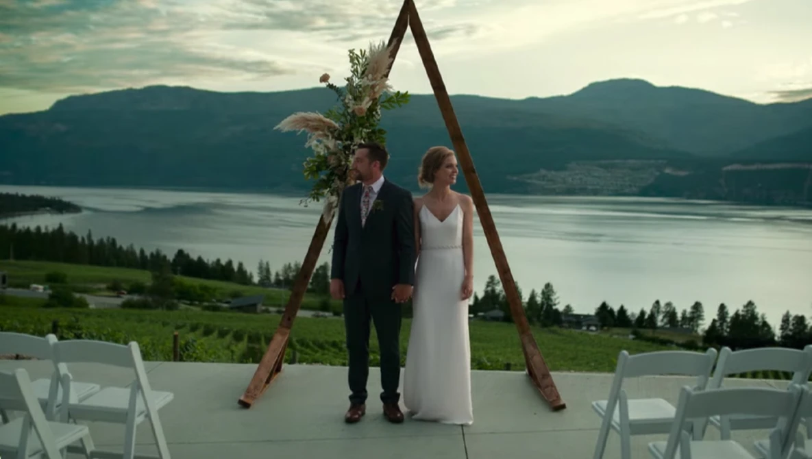 Bride and groom standing at wooden arch altar with lake and mountains in background.