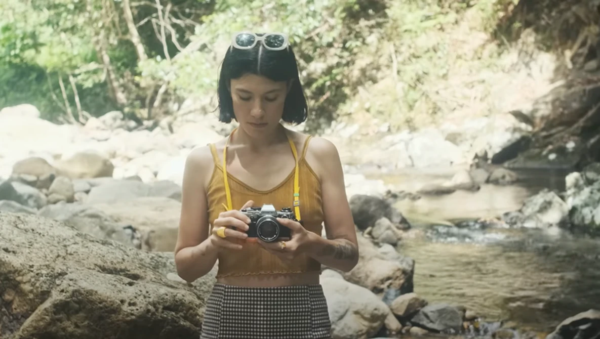 Woman in yellow top holding a vintage camera by a rocky stream.