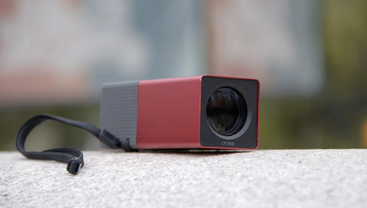 Compact red and gray projector with lens visible, sitting on concrete surface with blurred background.