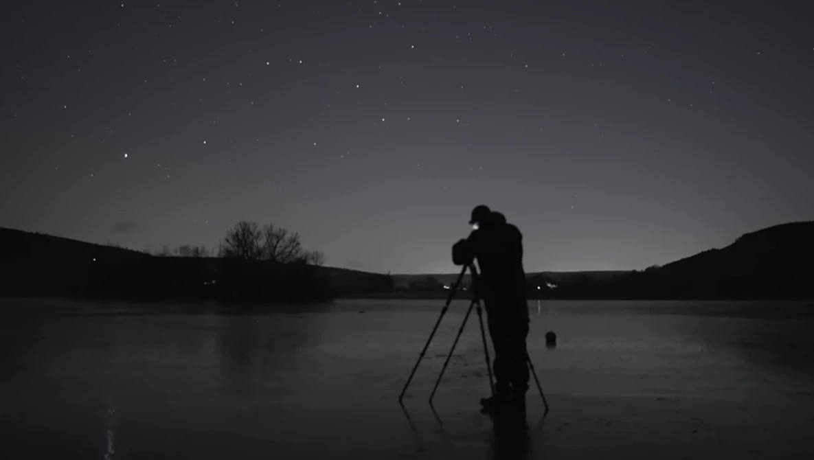 Silhouetted photographer on frozen lake at night with telescope on tripod pointed at starry sky.