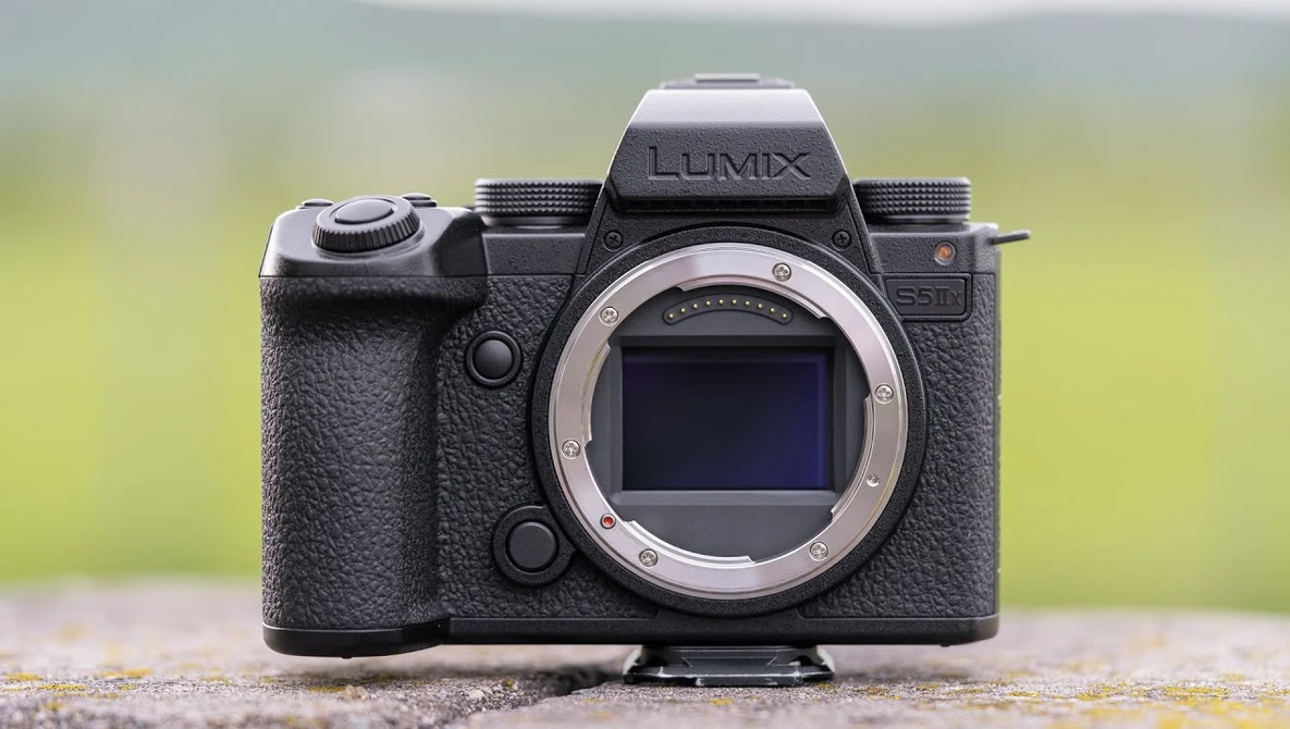 Panasonic Lumix mirrorless camera body with lens mount visible, positioned on a textured surface with blurred green background.