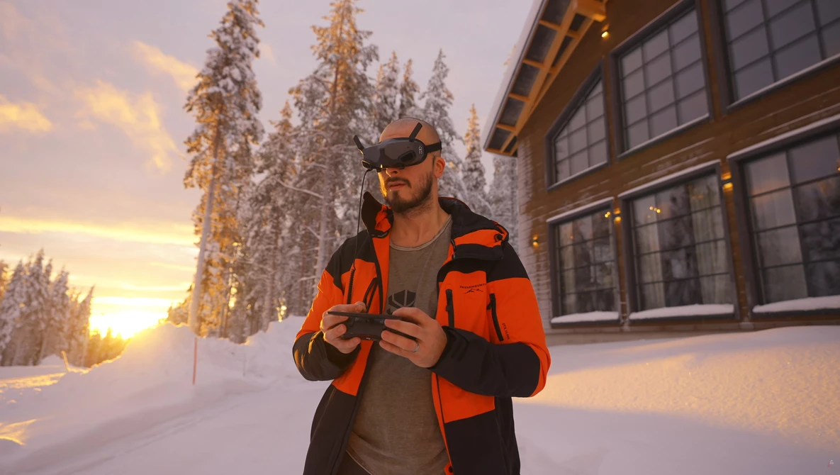 Man in bright orange jacket and dark cap standing in snowy landscape at golden hour.