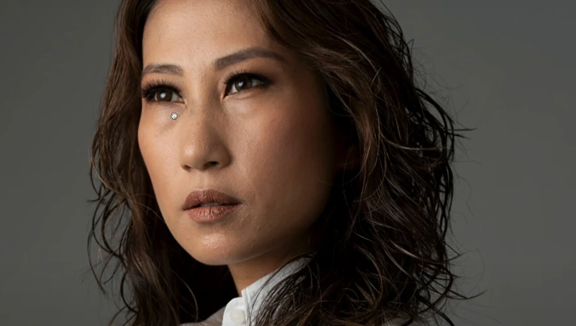 Portrait of a woman with wavy dark hair wearing a white shirt, gazing to the side against a neutral gray background.