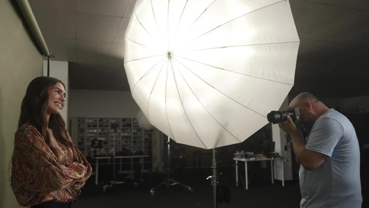 Photographer using a camera on tripod while model stands under large umbrella softbox in studio setting.