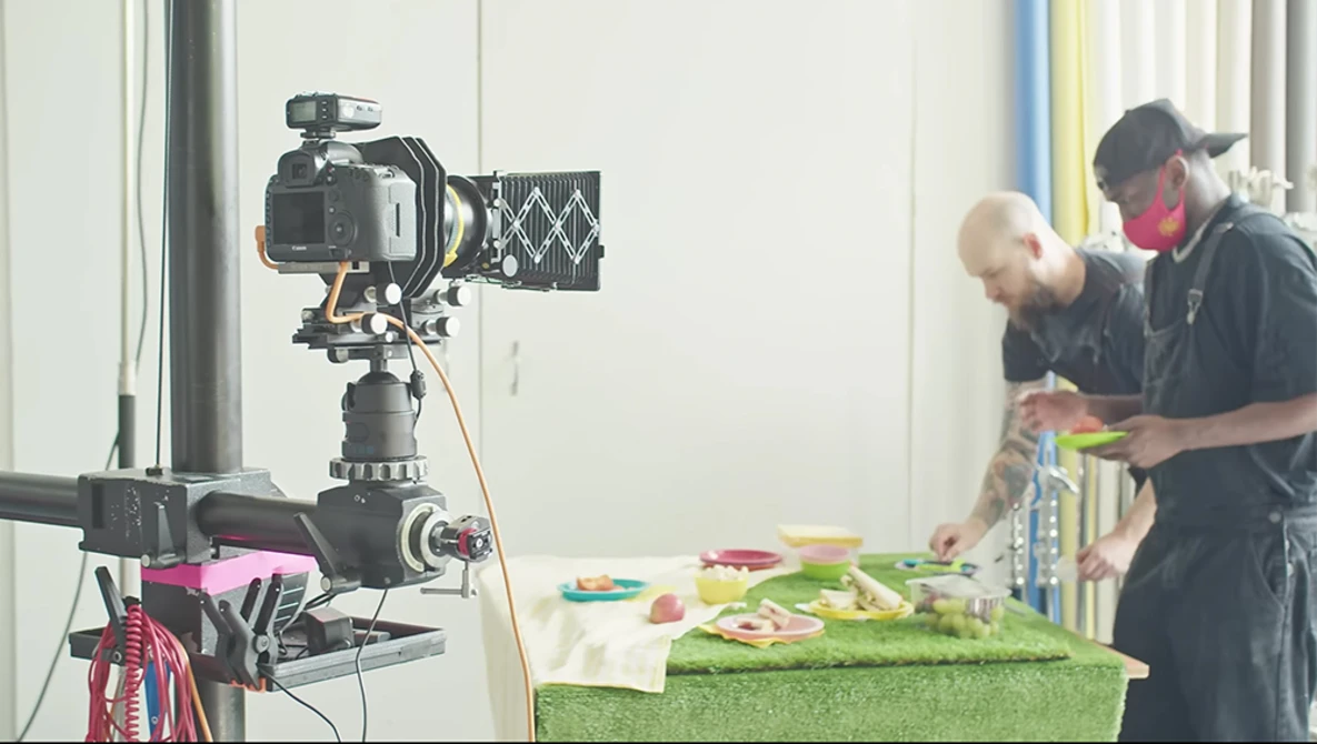 Professional video camera mounted on tripod filming two people preparing food at a table with green surface.