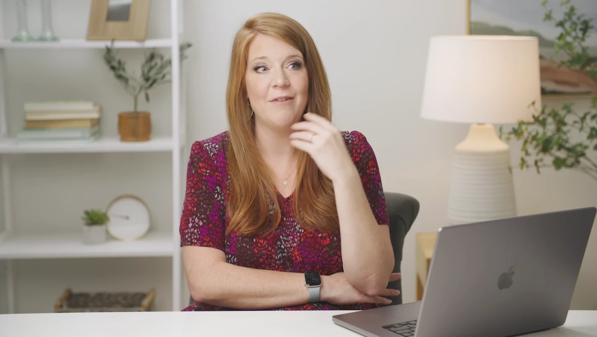 Woman with reddish-brown hair wearing a burgundy patterned shirt sits at a desk with a laptop, hand near her chin in a thoughtful pose.