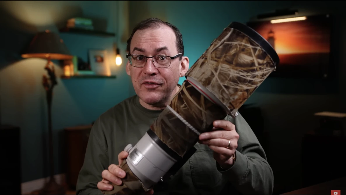 Man with glasses holding a camera lens and its protective case in a studio setting.