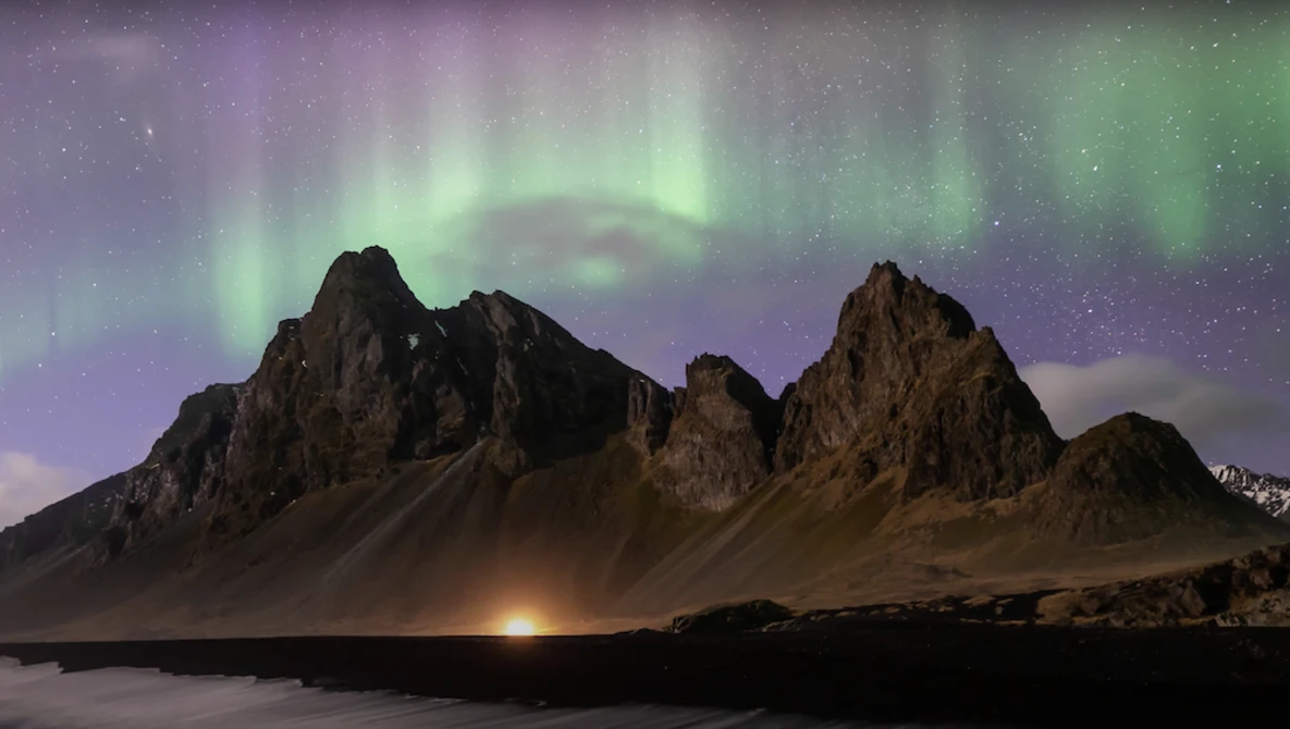 Jagged volcanic mountains silhouetted against a green and purple aurora borealis display over dark water.