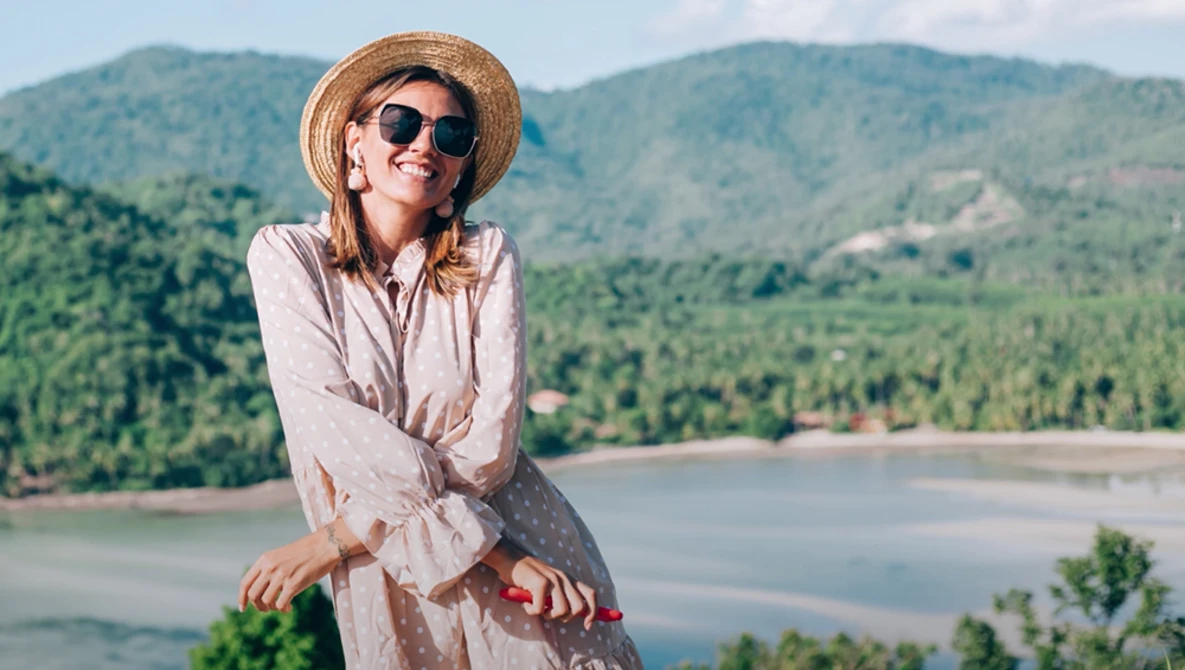 Woman in light shirt and straw hat smiling at camera overlooking forested mountains and river valley.