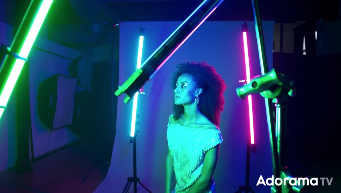 Woman posed among neon tube lights in cyan, green, pink, and blue creating a geometric light setup.