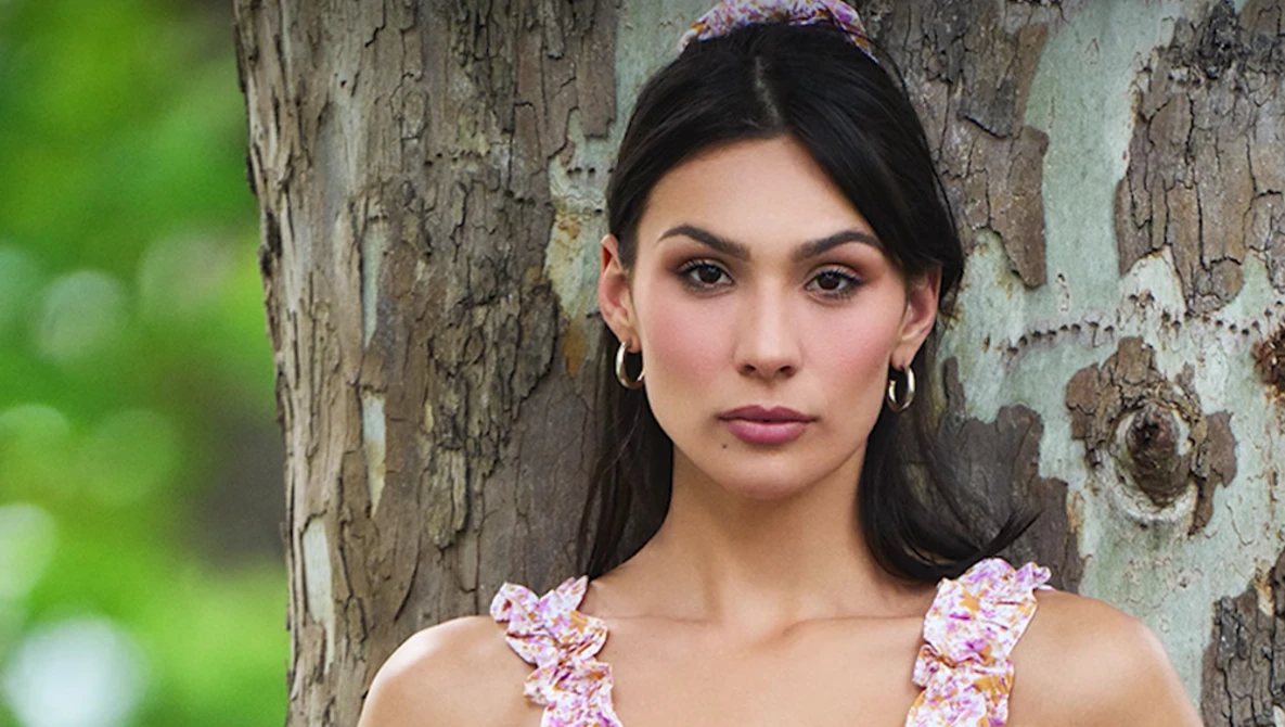 Woman with dark hair and hoop earrings leaning against weathered tree bark, wearing a floral patterned tank top.